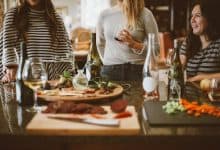 Three women standing around a table with food and wine.