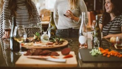 Three women standing around a table with food and wine.